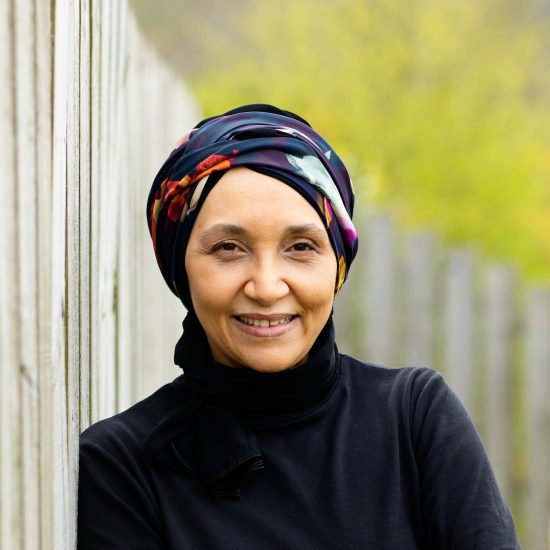 PEN Pinter Prize 2025 Winner, Leila Aboulela, dressed in a black top with a patterned headscarf and smiling at the camera, leaning against a wooden fence with greenery behind her.