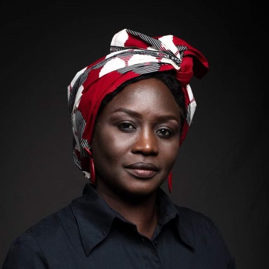 Stella Gaitano is wearing a red, white, and black patterned headscarf and a dark shirt and is posing against a dark background.