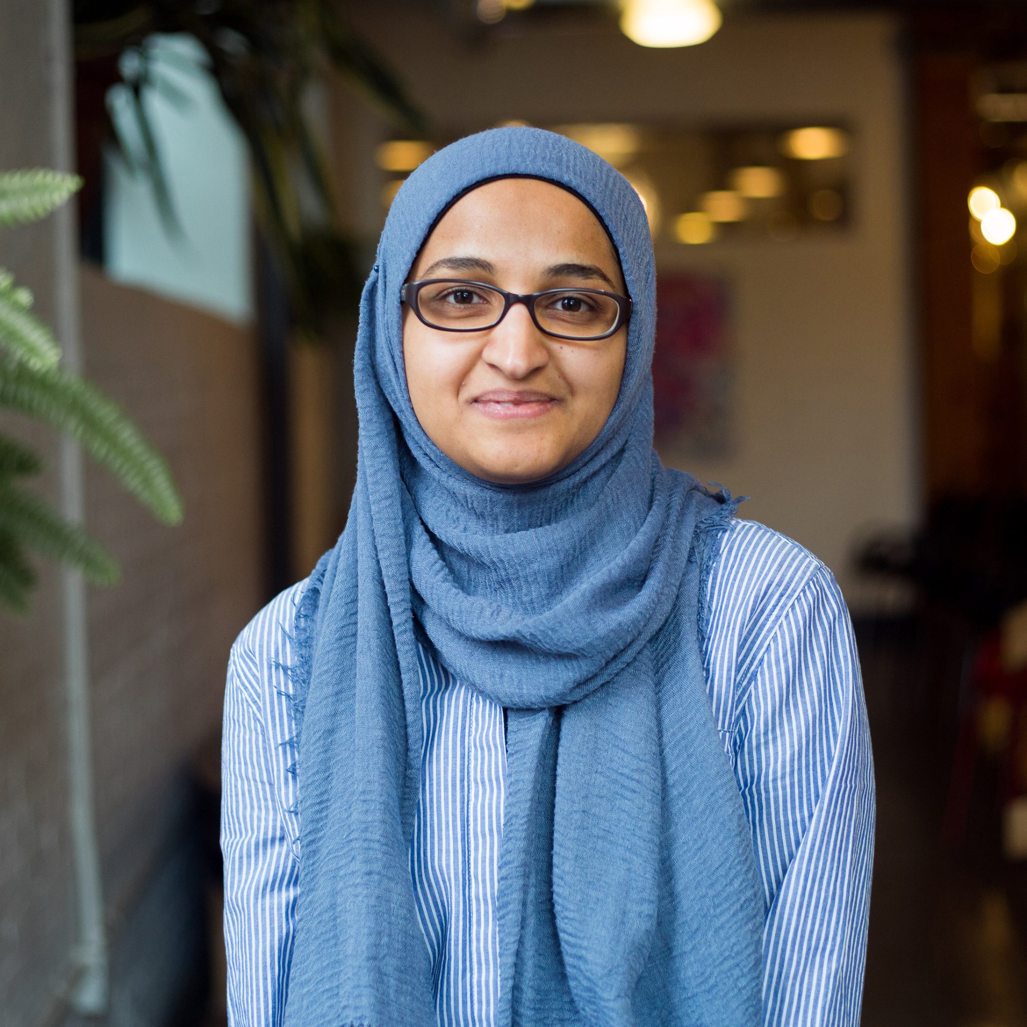 woman in glasses and a light blue head scarf standing in a softly lit space