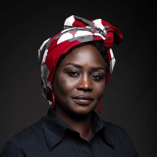 woman wearing a red, white, and black patterned headscarf and a dark shirt, posing against a dark background